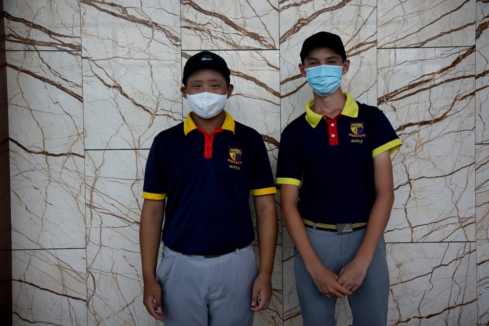 Two young males in matching navy and yellow golf polo shirts, black caps, and face masks stand before a marble wall.